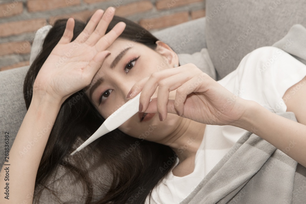 Naklejka premium Pretty Asian woman sick from cold and used with thermometer lying on sofa in the room.