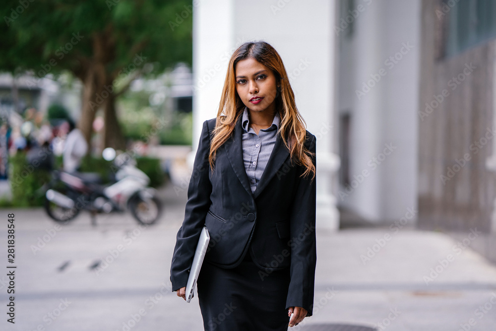 Portrait of a beautiful, young and attractive Singaporean Malay Asian woman in a business suit walking in a city in Asia during the day with her laptop computer. She is smiling confidently.