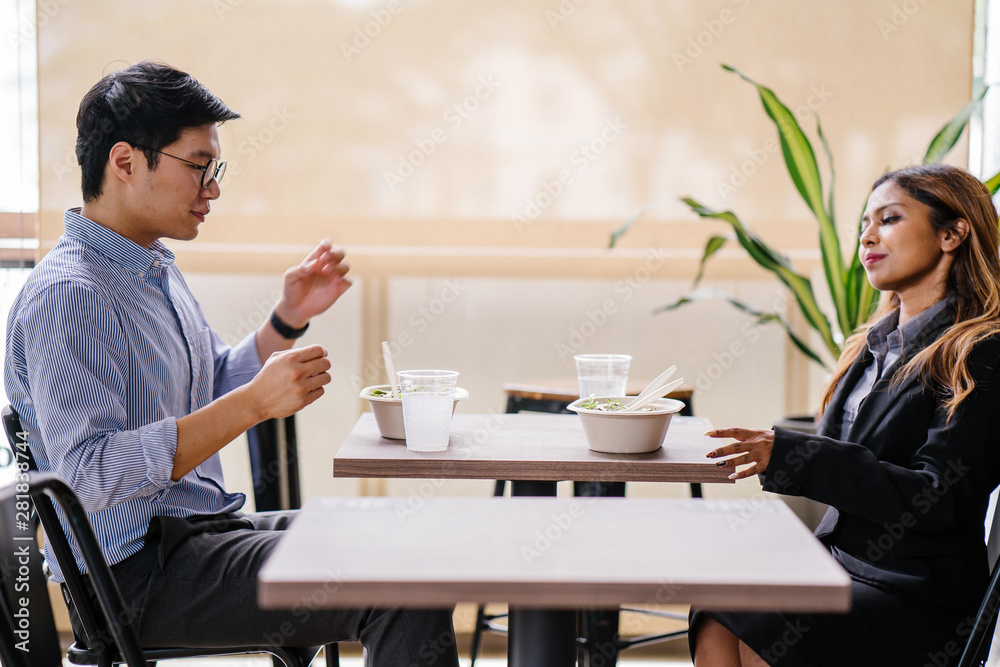 A pair of diverse colleagues sit and have a healthy salad lunch together. They are professionally dressed and chatting as they dig into their food. One is a Malay Asian woman, the other a Korean man. A pair of diverse colleagues sit and have a healthy salad lunch together. They are professionally dressed and chatting as they dig into their food. One is a Malay Asian woman, the other a Korean man.