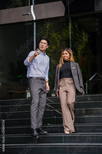 A tall, confident and clean-cut Asian man in shirt and pants smiles as he gives a tour or directions to the city to a young and beautiful Malay Asian woman in a suit during the day.