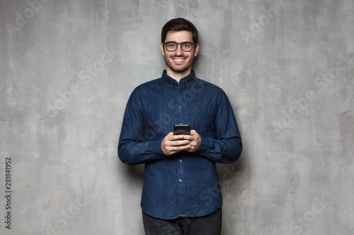 Smiling european man in denim shirt and trendy glasses standing against gray textured wall, holding his phone with both hands © Damir Khabirov