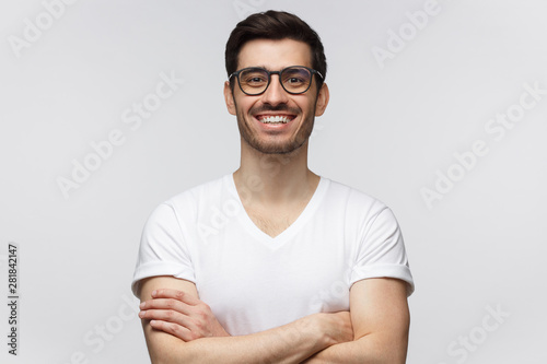 Cheerful confident young man in casual white t-shirt looking at camera, standing with crossed arms isolated on gray background