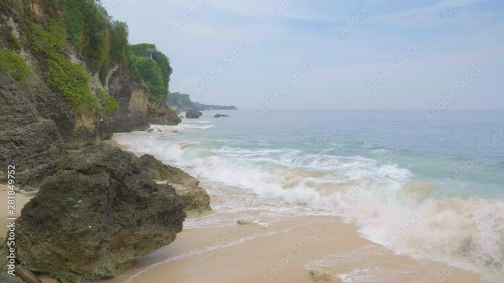 Slow motion of tropical beach with horizon in cloudy day with big waves in blue and green ocean and huge rock in the left Slow motion of tropical beach with horizon in cloudy day with big waves in blue and green ocean and huge rock in the left