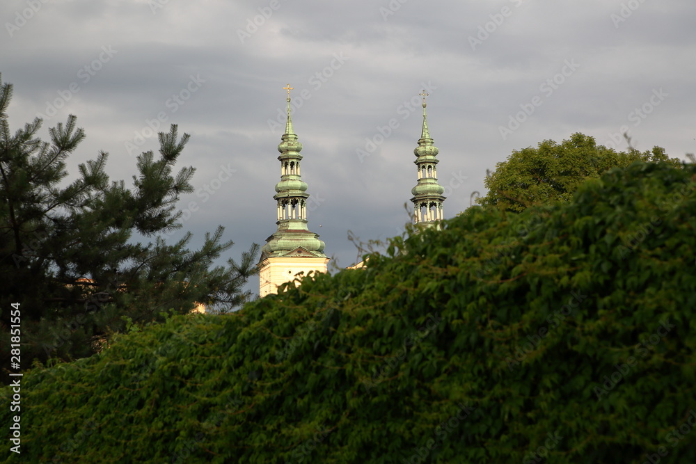 Towers of cathedral in Lowicz, Poland, visible from behind green thick bush, against cloudy sky