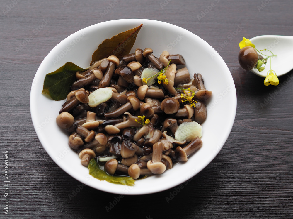 Delicious and beautiful pickled mushrooms and spices in a white plate, one fungus in a spoon on a wooden background. Home recipe pickles - small mushrooms with garlic and herbs close-up. Delicious and beautiful pickled mushrooms and spices in a white plate, one fungus in a spoon on a wooden background. Home recipe pickles - small mushrooms with garlic and herbs close-up.