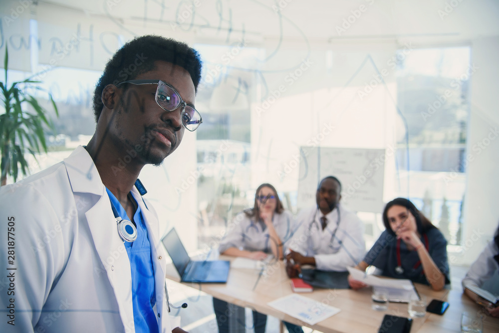 Fototapeta premium African professor at medical conference in modern clinic teaches his students. Doctor writing on board some formulas for interns in conference room at hospital at sunrise.