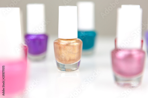 Table front view on a white desk full of small assorted glass and plastic bottles with nail polish and lacquer with focus on one golden bottle.