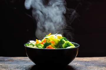The steam from the vegetables carrot broccoli Cauliflower in a black bowl, a steaming. Boiled hot Healthy food on table on black background,hot food and healthy meal concept