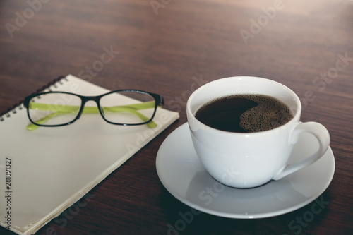 A cup of blackcoffee , blank notepad and eyeglasses on wooden table workpalce,