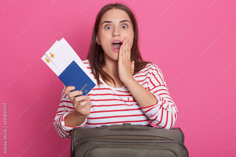 Photo of young astonished girl with dark straight hair dressed in casual shirt, posing with suitcase and holding passport with tickets in hands, looking at camera with open mouth over pink background.