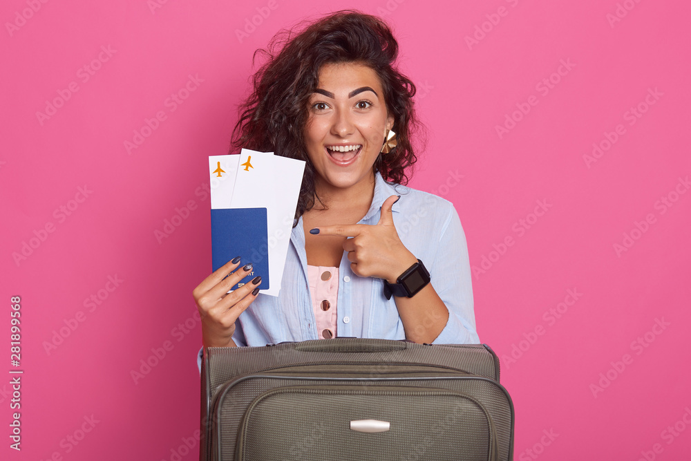 Studio shot of woman with suitcase, showing her tickets for plane in her hands with fore finger, isolated over rosy background, model with toothy smile, experesses happyness, wearinf stylish outfit. Studio shot of woman with suitcase, showing her tickets for plane in her hands with fore finger, isolated over rosy background, model with toothy smile, experesses happyness, wearinf stylish outfit.