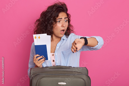 Close up of woman with dark curly hair, wearing fashionable outfit, stands with travel suitcase, holding documants in hands, late to trip, looks at watch with open mouth and shocking facial expression photo