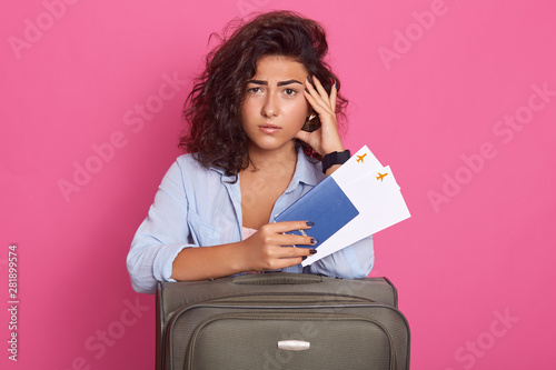 Photo of woman dressed fashionable clothes on pink background, looks tired and exausred, waiting her palne for long hours, posing with suitcase, passport and tickets. Traveling and lifestyle concept. photo