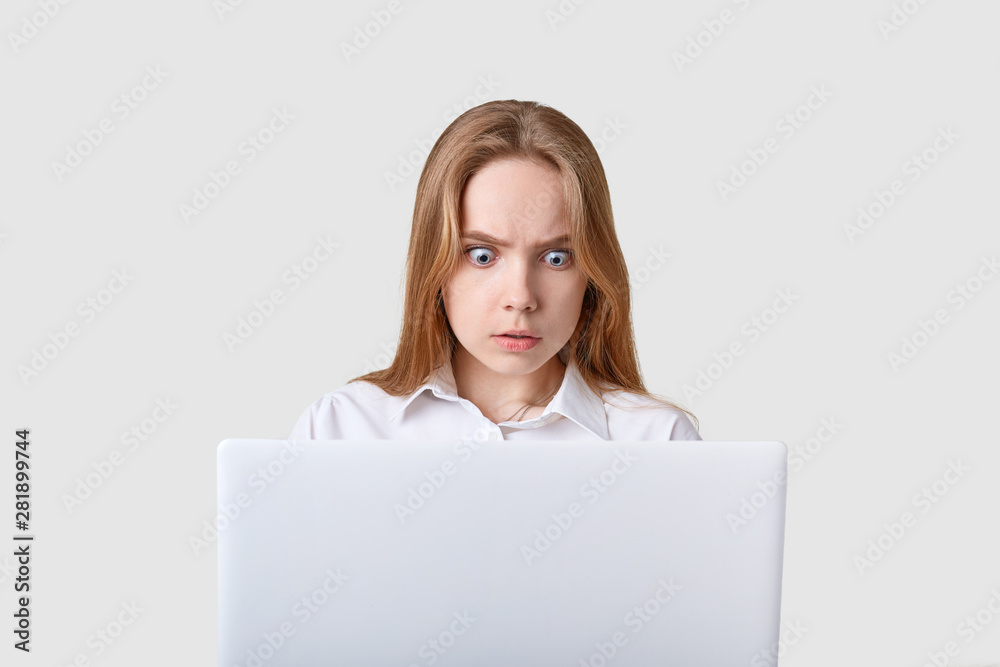 Portrait of shocked woman sitting in front of white laptop with wide opened eyes. Attractive girl looks at computer screen with astonished facial expression, sees somothing terrible, working online. Portrait of shocked woman sitting in front of white laptop with wide opened eyes. Attractive girl looks at computer screen with astonished facial expression, sees somothing terrible, working online.