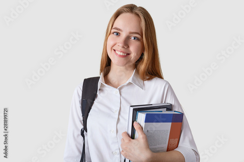 Indoor shot of beautiful teenager girl with black backpack and paper folder in hands, looking happy, attractive student woman isolated on white background. Teenagers, education and success concept. photo