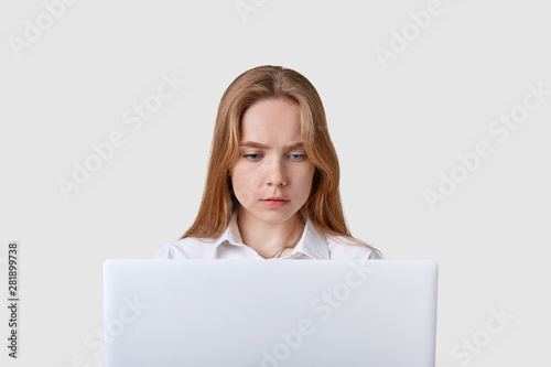 Front view of concentrated adult girl sitting in front of white portable laptop, having online work, looks at computer screen, wearing blouse, has straight fair hair, using wireless Internet.