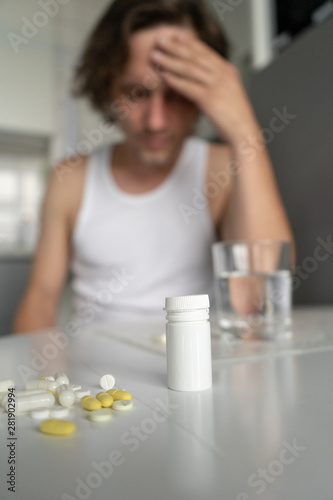 Pills and tablets lying on the table. A man holds his hand on his head. Sickness, malaise, headache, cold. Foreground. Vertical. Close-up