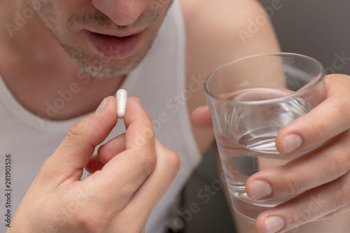 A man holds a pill and a glass of water, going to drink the medicine. Sickness, malaise, headache, cold. Foreground. Front view. Close-up