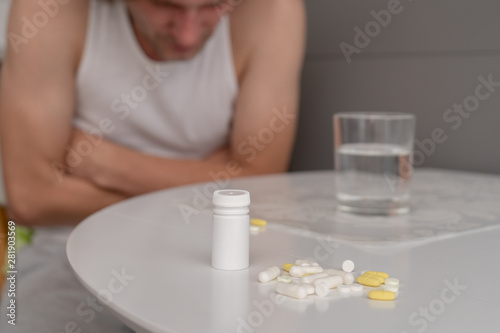 A man in home clothes sits on a chair and holds his hands on his stomach at the white kitchen table, on which there are tablets, pills and a glass of water. Severe abdominal pain, malaise. Close-up