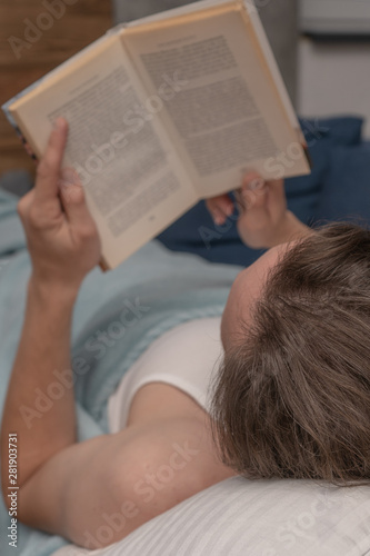 A man lies at home on the sofa ander the blanket and read book. Education, studying, View from the left