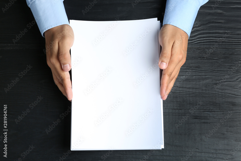Man holding blank paper sheets for brochure at black wooden table, top view. Mock up Man holding blank paper sheets for brochure at black wooden table, top view. Mock up