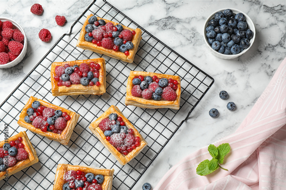 Cooling rack and fresh delicious puff pastry with sweet berries on white marble table, flat lay