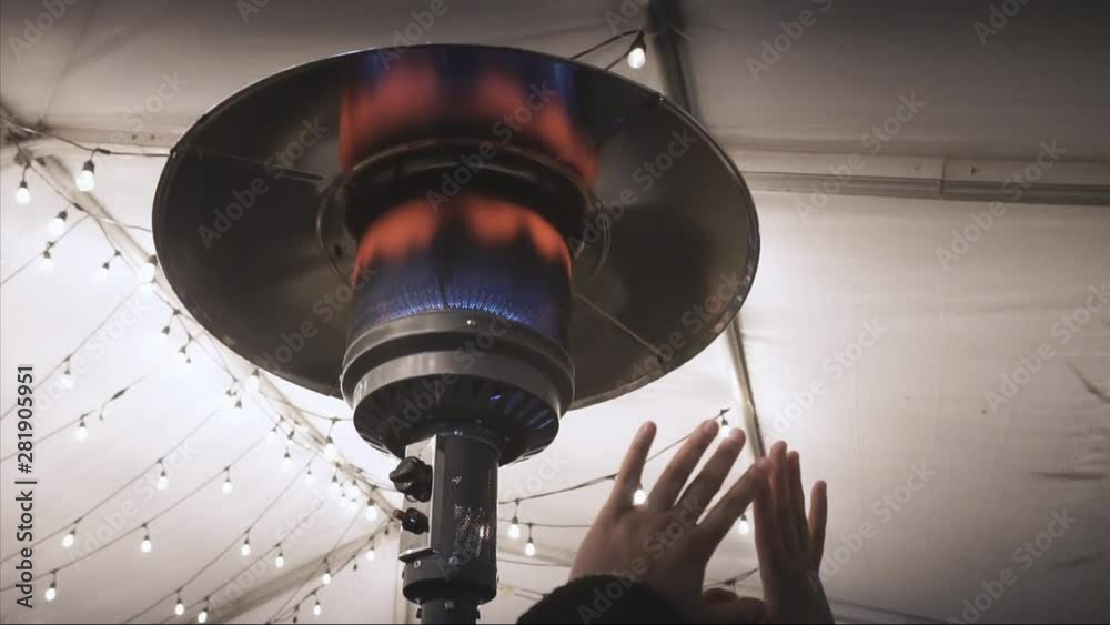 Woman is warming her hands near an artisan winter gas lamp fireplace or kerosene lamp outdoors, Decorative restaurant gas flame outdoor with led Christmas lights on the background. Slow motion.