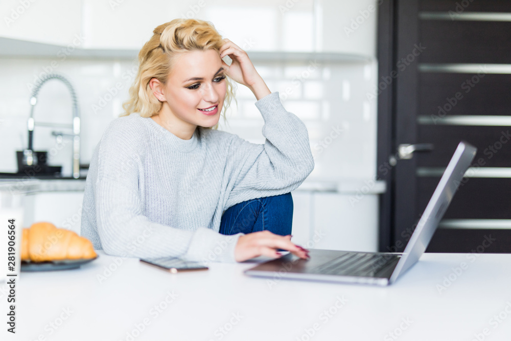 Smiling blonde woman having breakfast and using her laptop looking at the camera in the kitchen Smiling blonde woman having breakfast and using her laptop looking at the camera in the kitchen