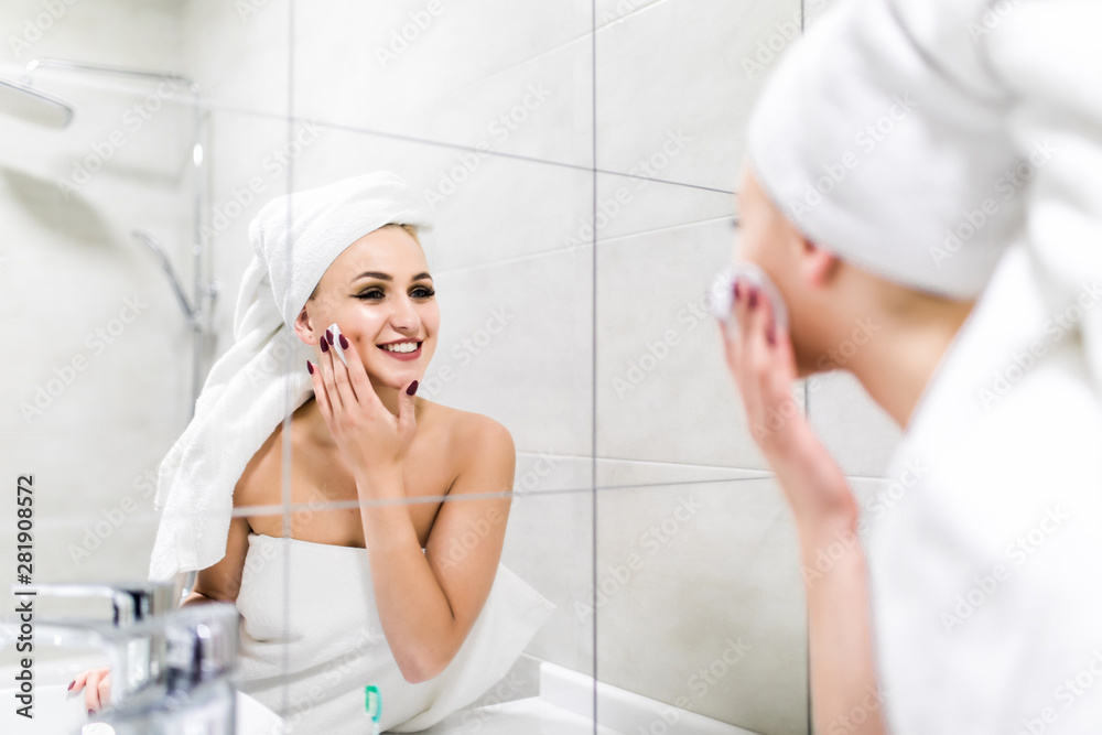 Young attractive woman looking at mirror, cleanses the face with a cotton pad after a shower at the bathroom. Skincare concept. Young attractive woman looking at mirror, cleanses the face with a cotton pad after a shower at the bathroom. Skincare concept.
