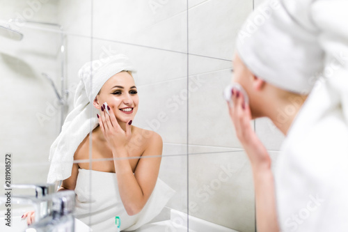 Young attractive woman looking at mirror, cleanses the face with a cotton pad after a shower at the bathroom. Skincare concept.