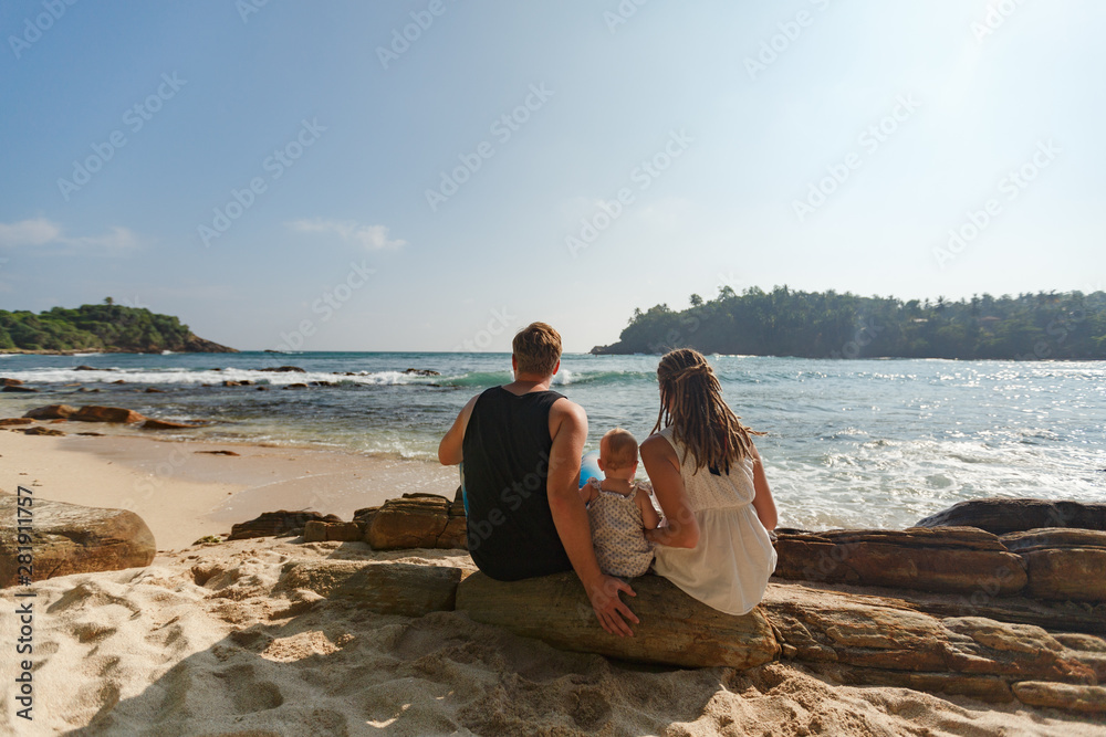 Back view of family with little child sitting on tropical sand beach, mother with dreads, father and baby, enjoying vacation. Vacation with kids