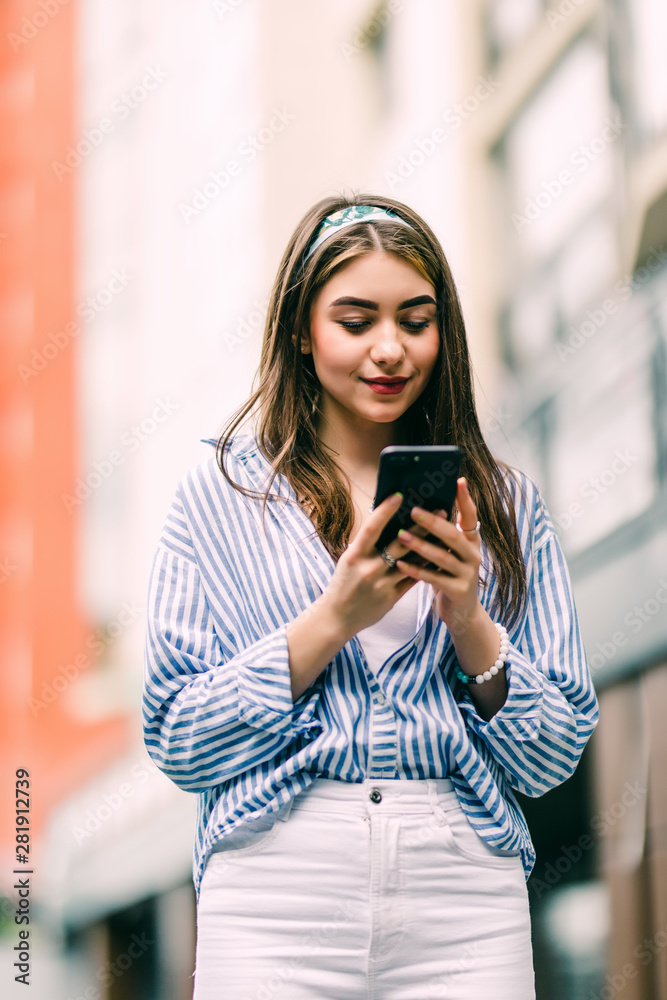 Fototapeta premium Young woman walking and using a smart phone in the street in a sunny summer day