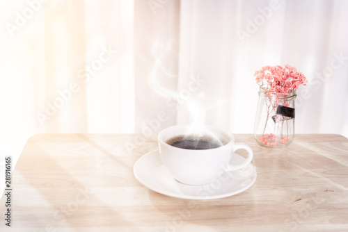 White coffee cup with smoke, red dried flower on wooden office desk  with morning sunlight and  soft curtain background