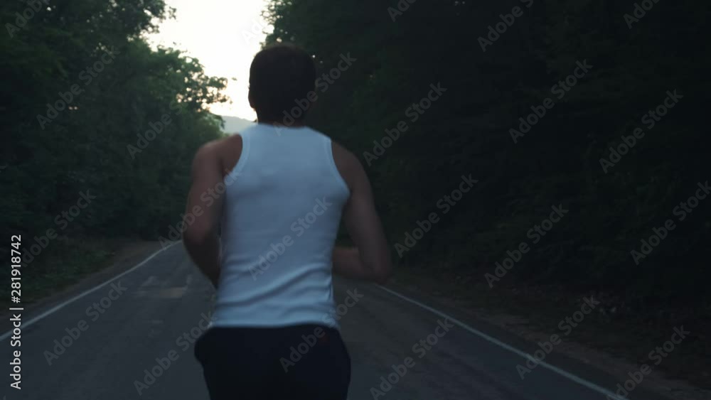 Man running outside in nature at rainy evening. Male runner doing a run at dusk on a highway among pines in the forest. Engaged in an active sports lifestyle. Strengthens your body. Sports in nature.