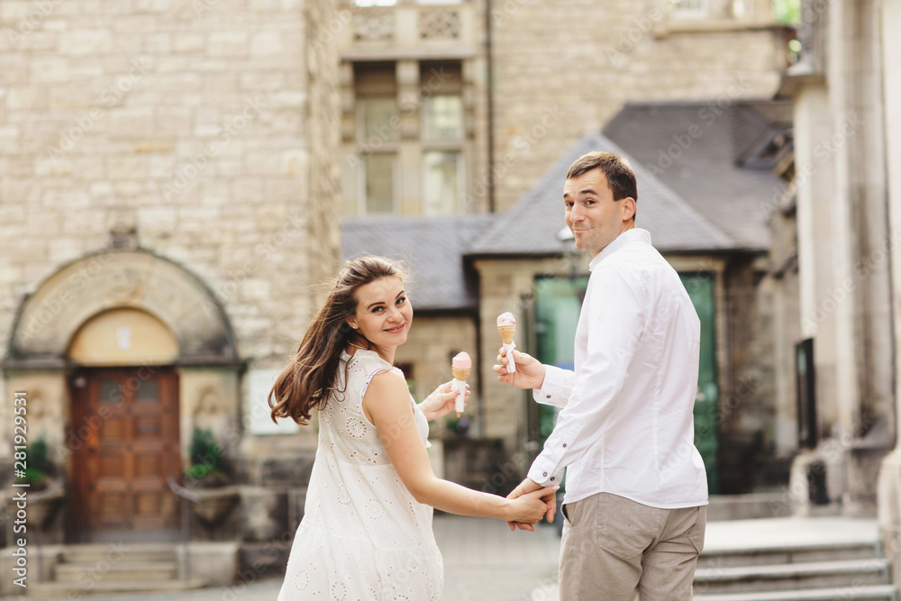 Pregnant woman in dress and her husband are walking in the city. Smiling couple enjoys a walk and eats ice cream. Back view