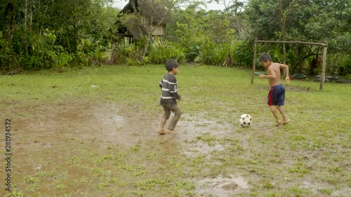 Two Young Indigenous Boys Are Playing Football On A Muddy Pitch Somewhere Outside In Their Rural Village Older Boy Scores A Goal General Shot, Camera Follow Up, Camera Movement In Ecuador