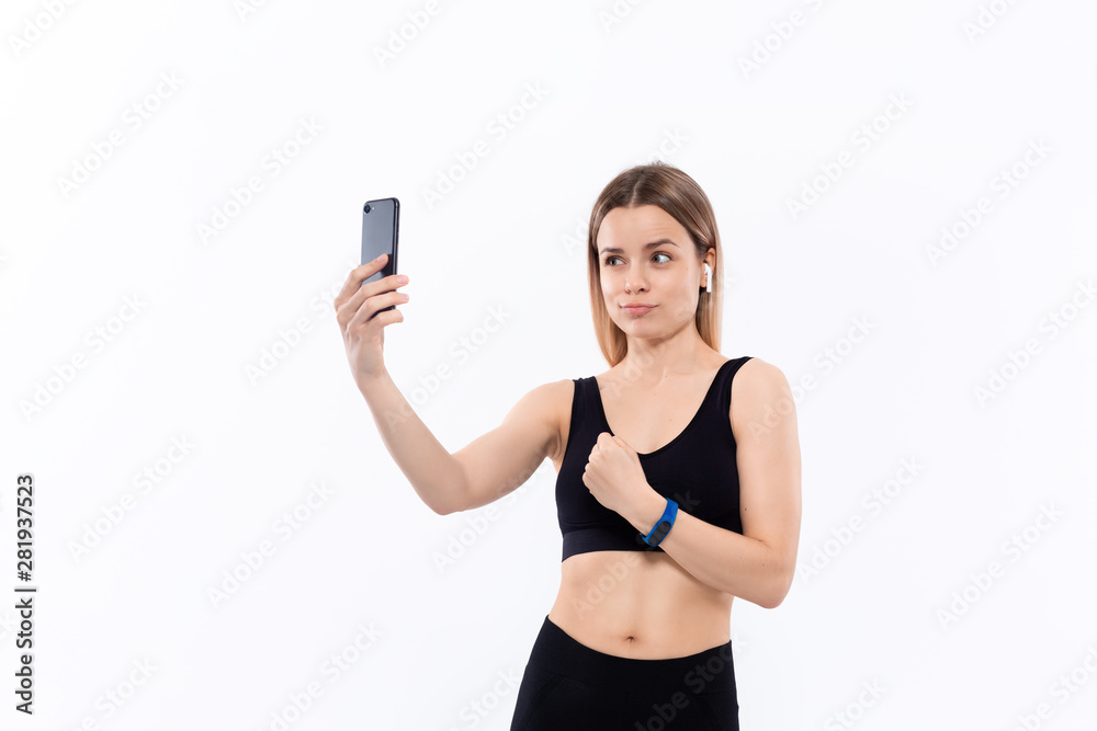 Young sporty blond woman in a black sportswear making selfie with smartphone showing bicep after workout standing over white background. Model listens music during workout with wireless headphones Young sporty blond woman in a black sportswear making selfie with smartphone showing bicep after workout standing over white background. Model listens music during workout with wireless headphones