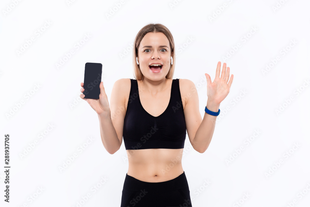 Excited young sporty blond woman in a black sportswear with wireless ear buds showing screen of a smartphone with a finger standing over white background. Excited young sporty blond woman in a black sportswear with wireless ear buds showing screen of a smartphone with a finger standing over white background.