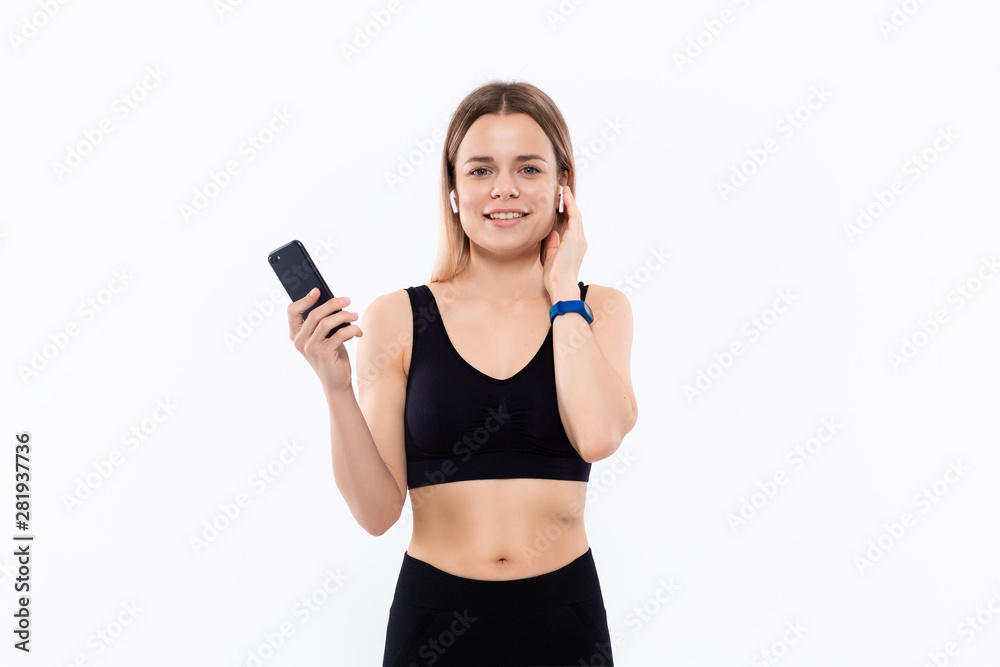 Excited young sporty blond woman in a black sportswear with wireless ear buds looking at the screen of a smartphone standing isolated over white background. Excited young sporty blond woman in a black sportswear with wireless ear buds looking at the screen of a smartphone standing isolated over white background.