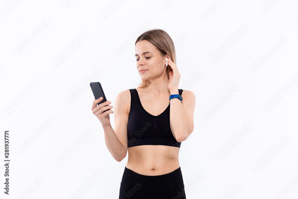 Excited young sporty blond woman in a black sportswear with wireless ear buds looking at the screen of a smartphone standing isolated over white background. Excited young sporty blond woman in a black sportswear with wireless ear buds looking at the screen of a smartphone standing isolated over white background.