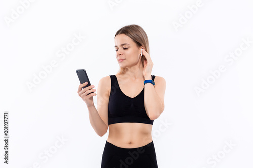 Excited young sporty blond woman in a black sportswear with wireless ear buds looking at the screen of a smartphone standing isolated over white background.