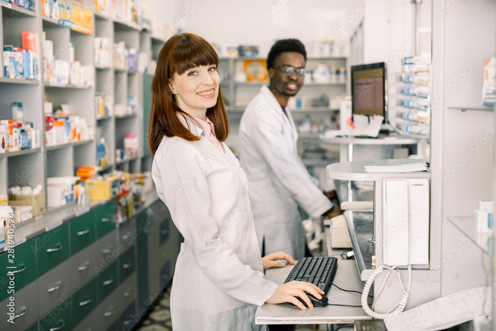 Two young cheerful pharmacists African man and Caucasian woman working together and using computers. Pharmacists searching medicines at pharmacy