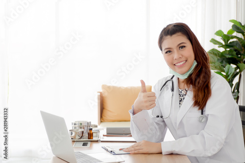 Smiling young female physician wear white coat with hand thumbs up using laptop while working at examination room, healthcare and medical concept.