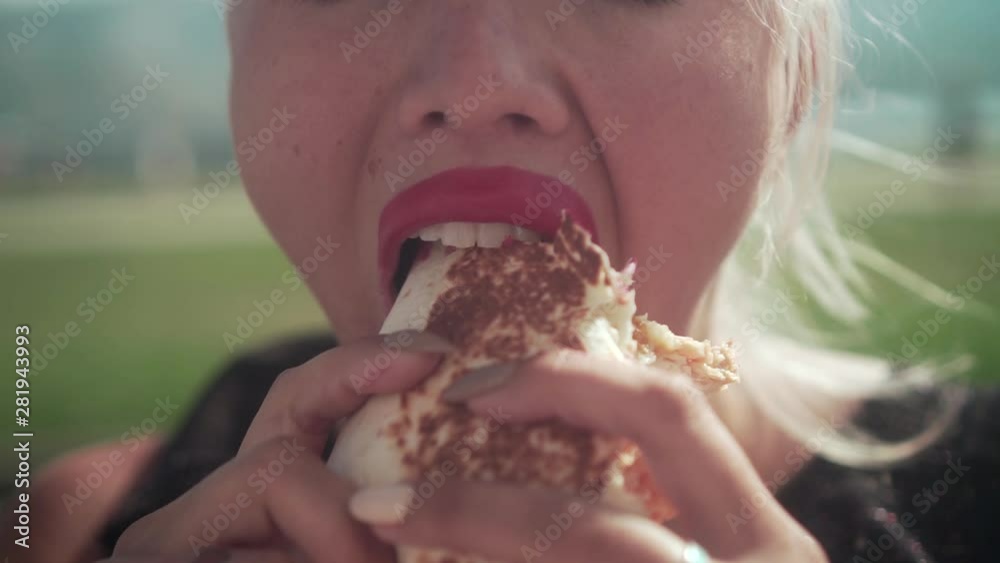 A hungry blonde woman eating a hamburger on the street. Young woman with unevenly painted lips eagerly eat a hamburger, holding in their hands. Unhealthy diet, fast food concept.