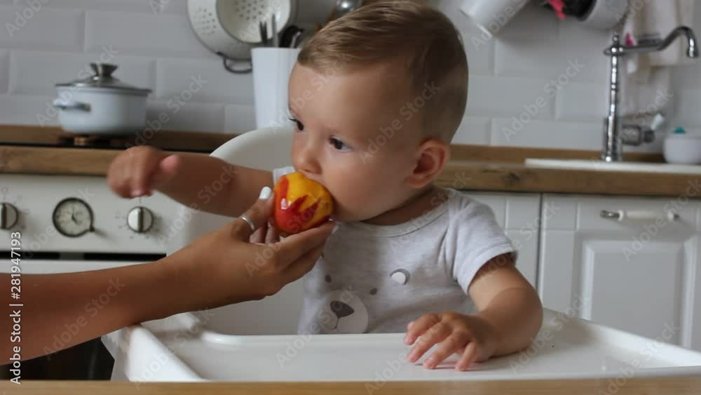 Charming little baby boy eating first food peach at the kitchen. Mom feeds child Charming little baby boy eating first food peach at the kitchen. Mom feeds child