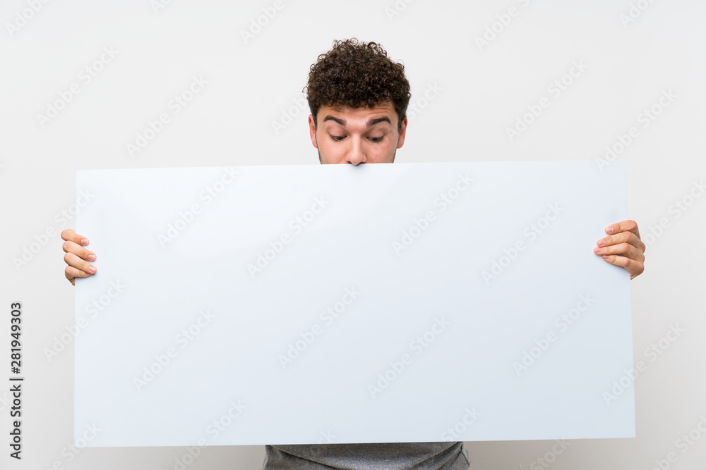 Man with curly hair over isolated wall holding an empty white placard for insert a concept Man with curly hair over isolated wall holding an empty white placard for insert a concept
