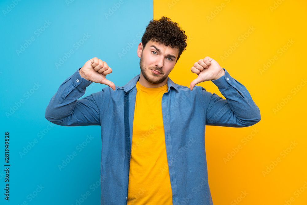 Man with curly hair over colorful wall showing thumb down Man with curly hair over colorful wall showing thumb down
