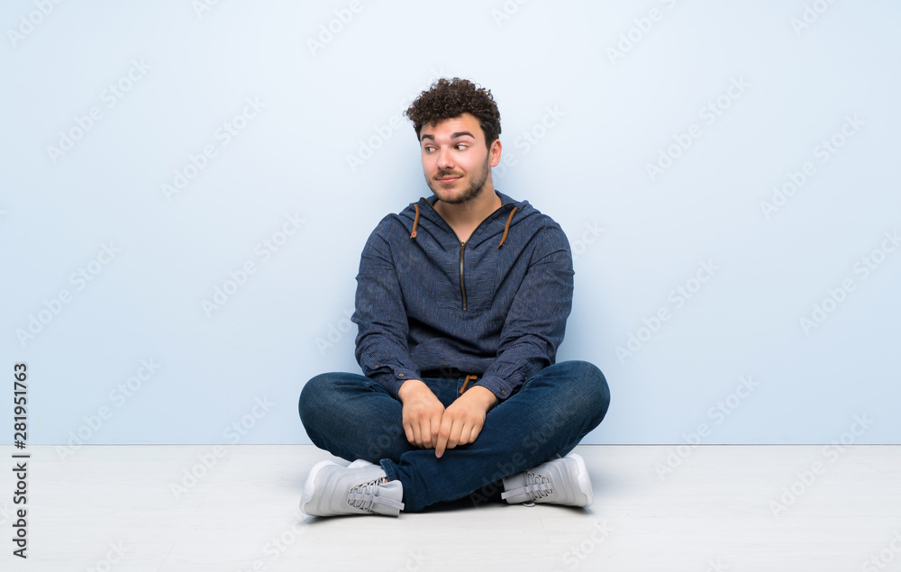 Young man sitting on the floor making doubts gesture looking side Young man sitting on the floor making doubts gesture looking side