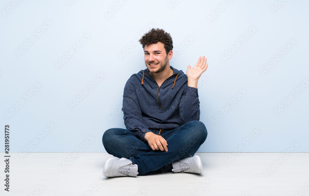 Young man sitting on the floor saluting with hand with happy expression
