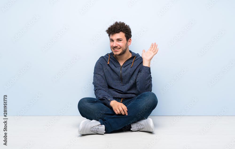 Young man sitting on the floor saluting with hand with happy expression Young man sitting on the floor saluting with hand with happy expression